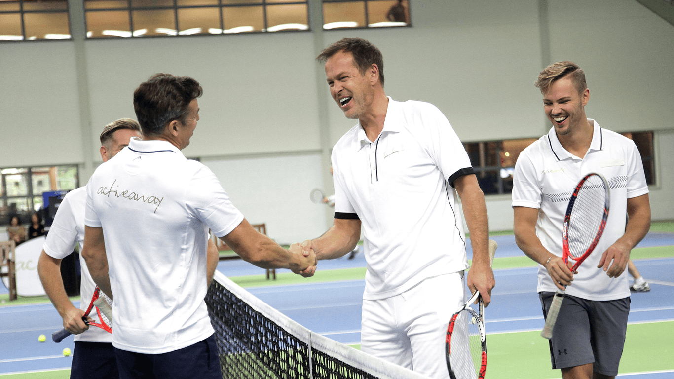 Men shake hands at the net after an indoor tennis match, smiling with rackets.