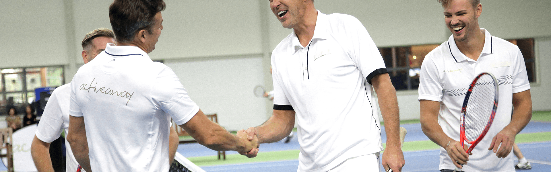 Men shake hands at the net after an indoor tennis match, smiling with rackets.