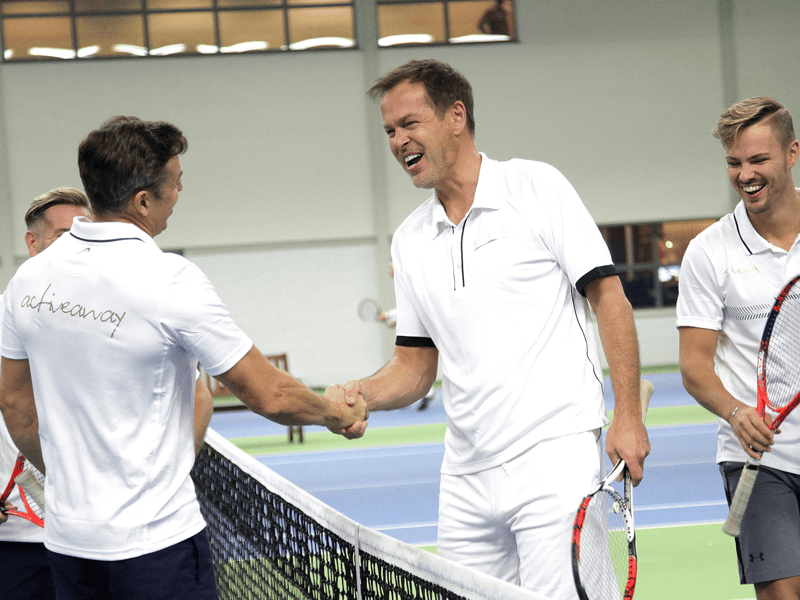 Men shake hands at the net after an indoor tennis match, smiling with rackets.