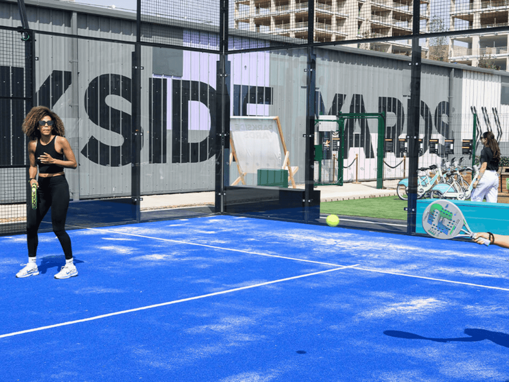 Players rally on a bright blue outdoor padel court as the ball flies across.