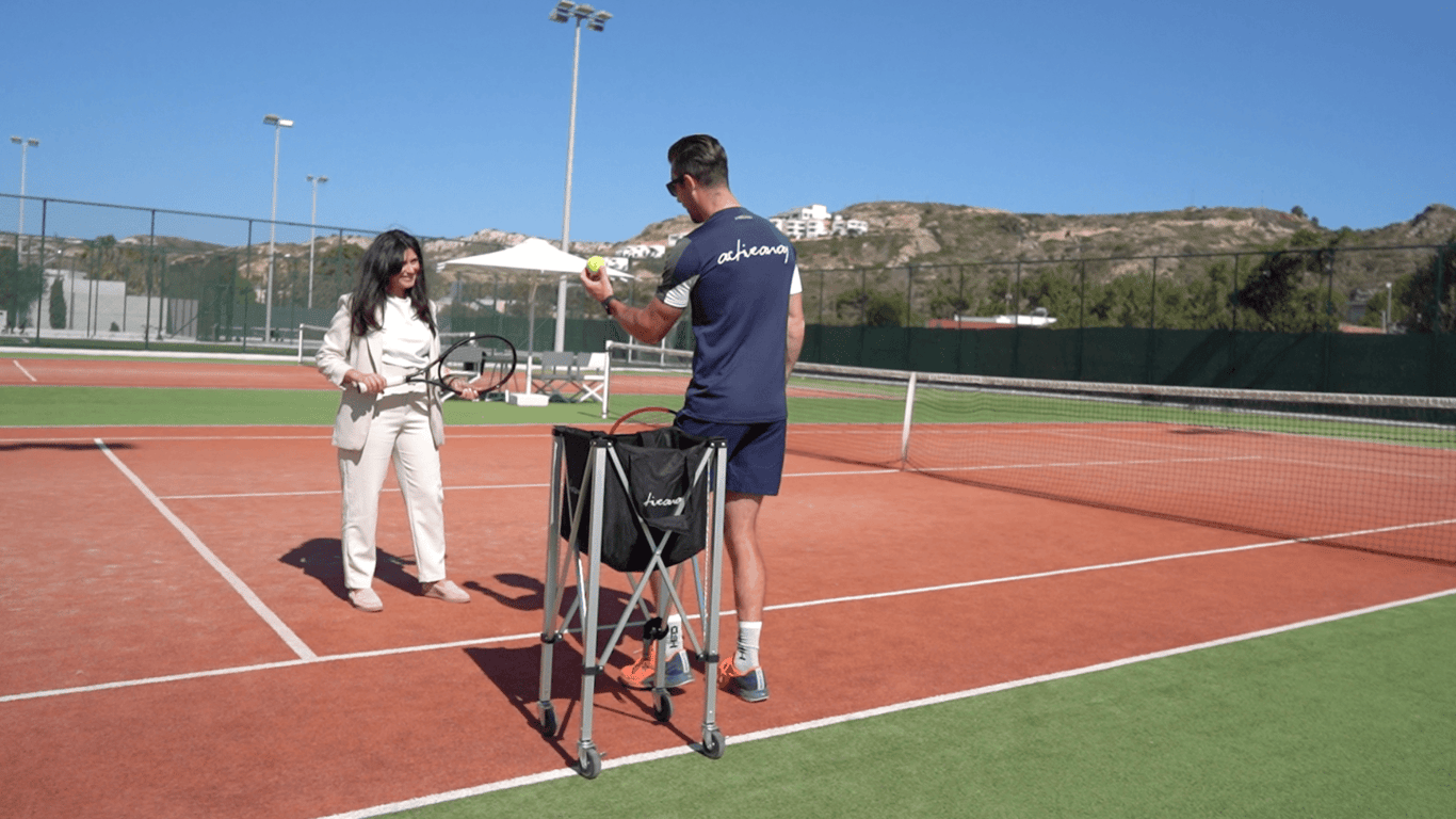 Tennis coach showing a drill to a guest beside a ball basket on a sunny court at a resort.