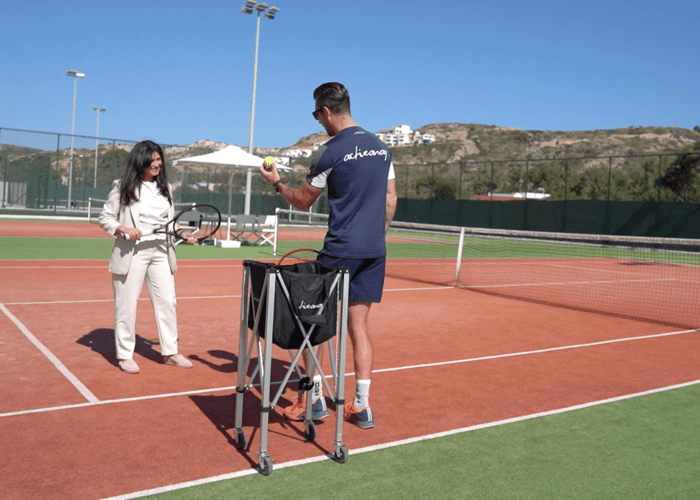Tennis coach showing a drill to a guest beside a ball basket on a sunny court at a resort.