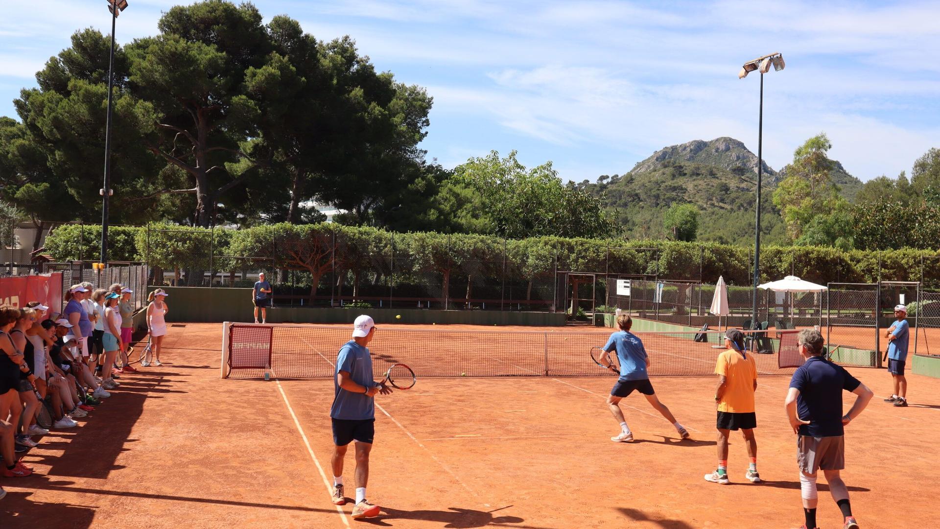 Group tennis clinic on a clay court, players rally at a mini-net as others watch, trees and hills in background.