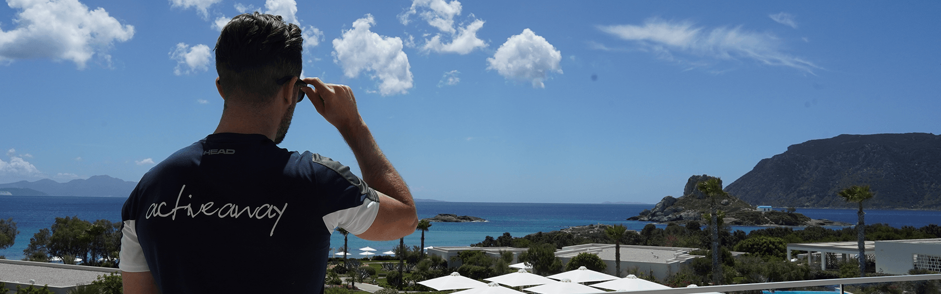 Man in Active Away shirt looking over a seaside resort with pool and mountains on a sunny day.