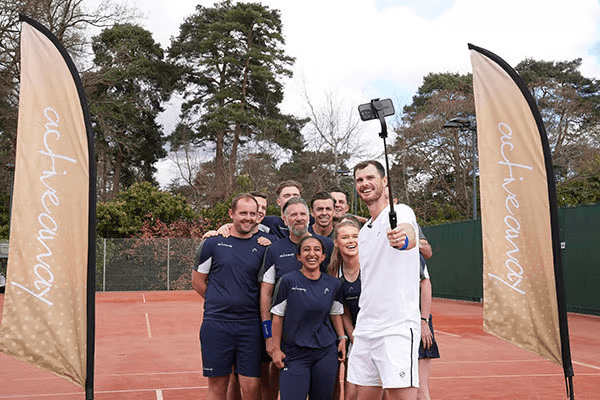 Tennis group selfie on a clay court, a player holds a selfie stick between two Active Away flags.