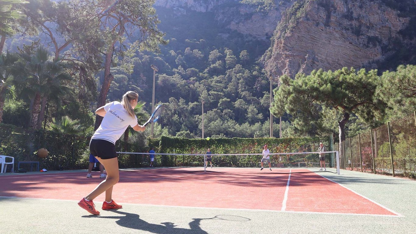 Woman hitting a tennis shot on a sunlit outdoor court with mountains and trees in the background.