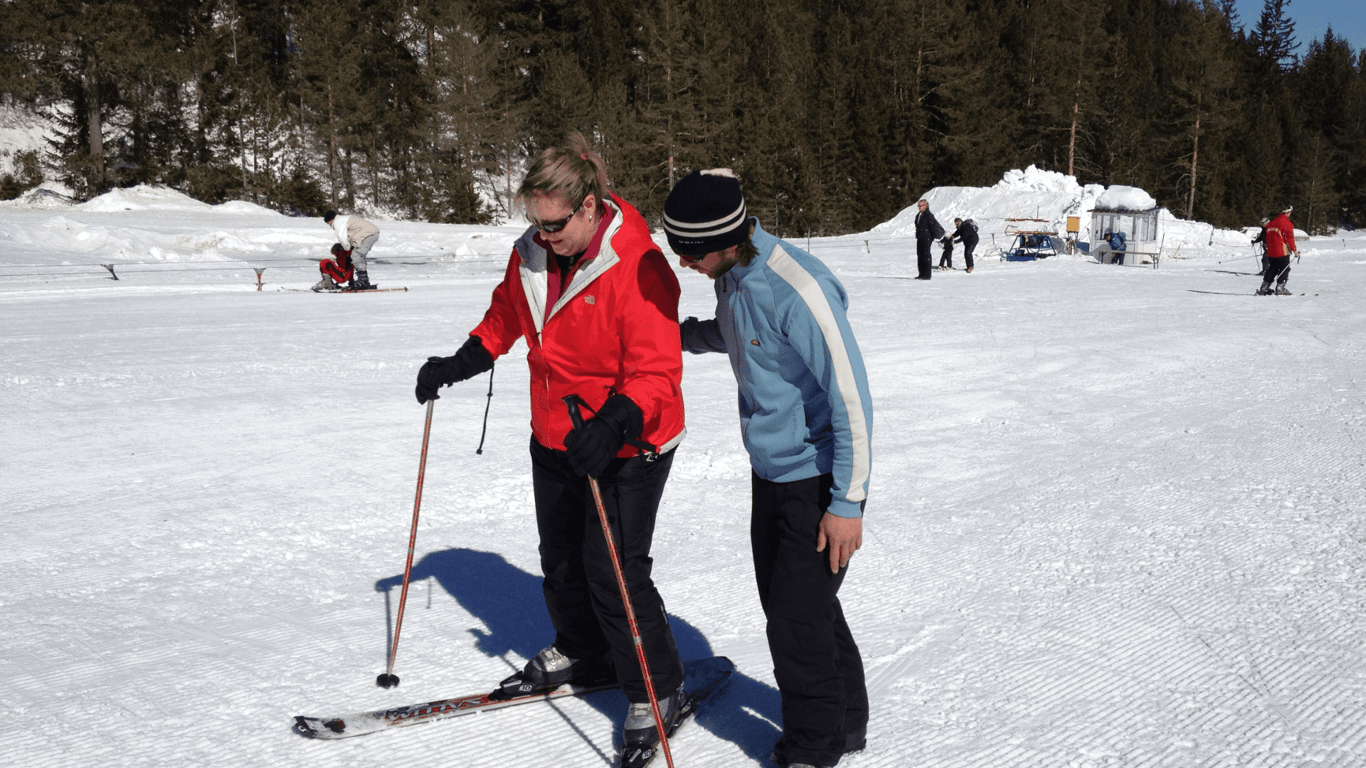 Ski instructor guiding a guest on a sunny slope, both in winter gear with poles.