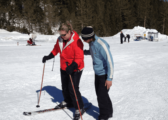 Ski instructor guiding a guest on a sunny slope, both in winter gear with poles.