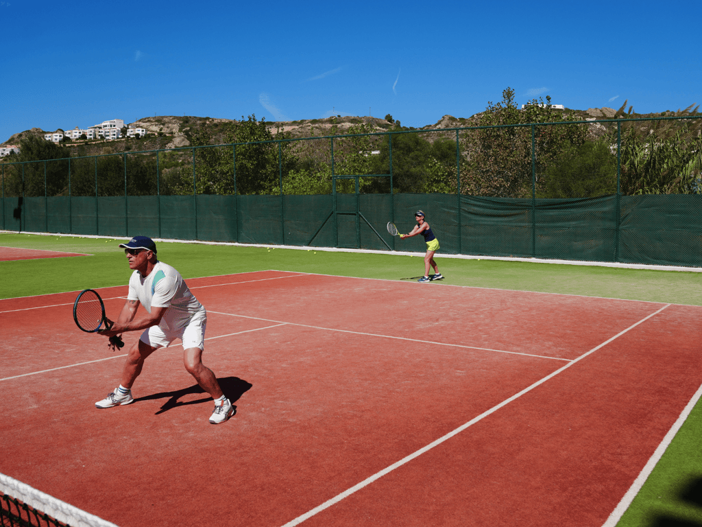 Two players practising doubles on a sunny outdoor tennis court, one at the net and one preparing to return.