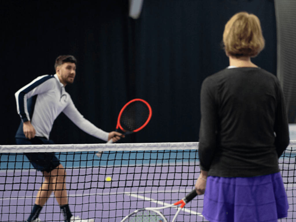 Two people rally on an indoor tennis court near the net, practising volleys.