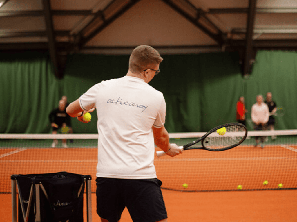 Tennis coach feeds balls during an indoor lesson, players ready across the net.