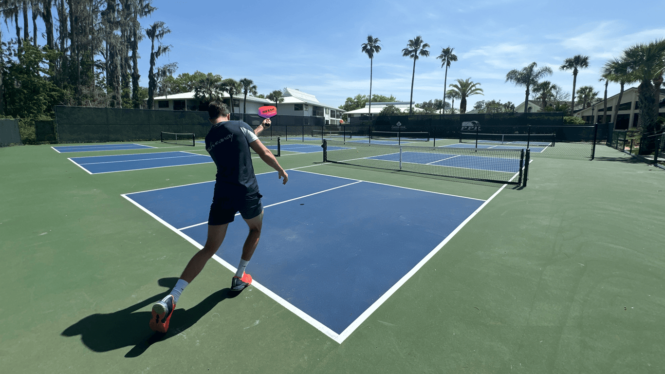 Player hitting a forehand on an outdoor pickleball court under a sunny sky with palm trees.