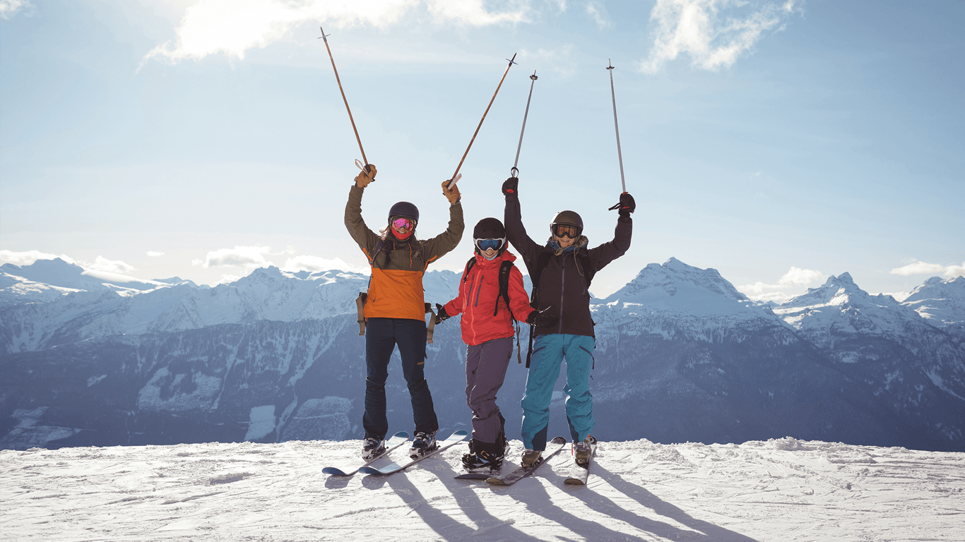 Three snowboarders raise ski poles on a sunny mountain summit with snowy peaks behind.