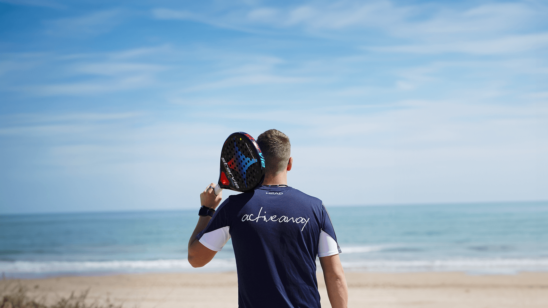 Padel player on a beach holding a racket over shoulder, wearing an Active Away shirt.