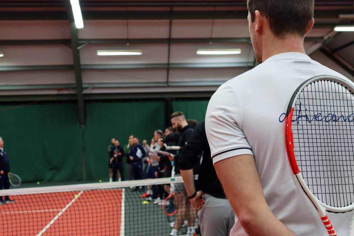 Tennis coach in a white shirt holding a racket stands by the net on a red indoor court, observing a group coaching session.