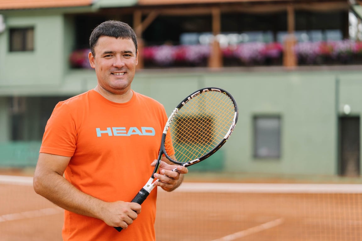 Smiling tennis coach in an orange shirt holding a racquet on a clay court.