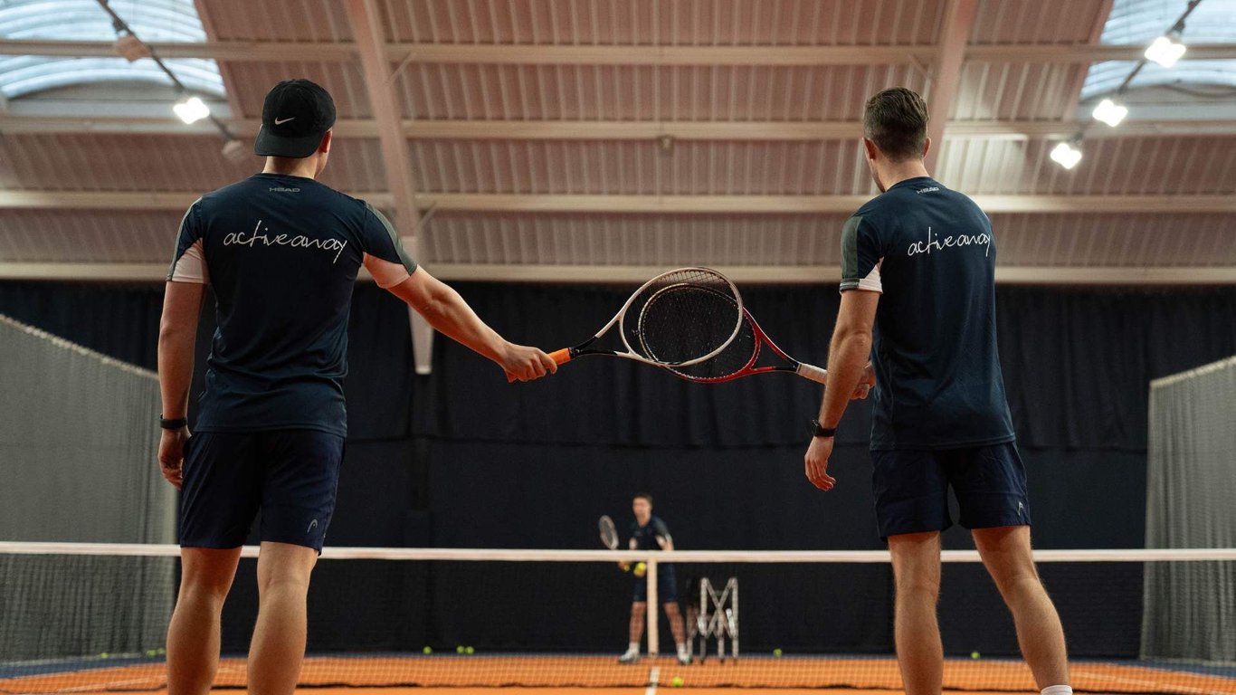 Two tennis players tap racquets on an indoor court, with a coach and balls visible in the background.