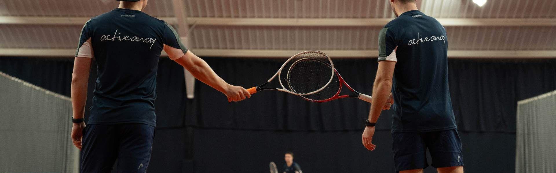 Two tennis players tap racquets on an indoor court, with a coach and balls visible in the background.