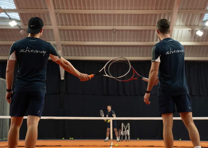 Two tennis players tap racquets on an indoor court, with a coach and balls visible in the background.