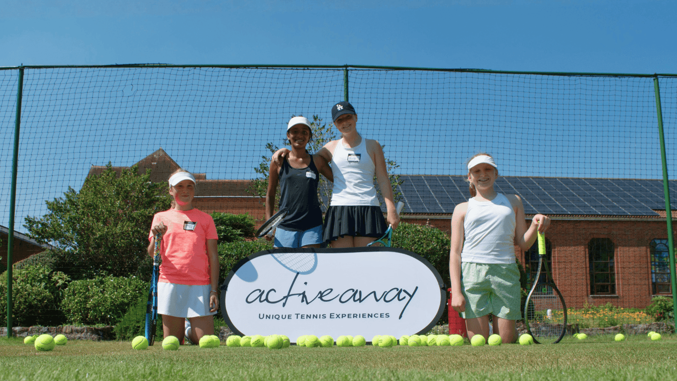 Four junior tennis players smiling on a grass court with rackets and an Active Away sign.