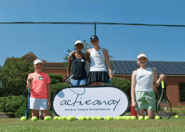 Four junior tennis players smiling on a grass court with rackets and an Active Away sign.