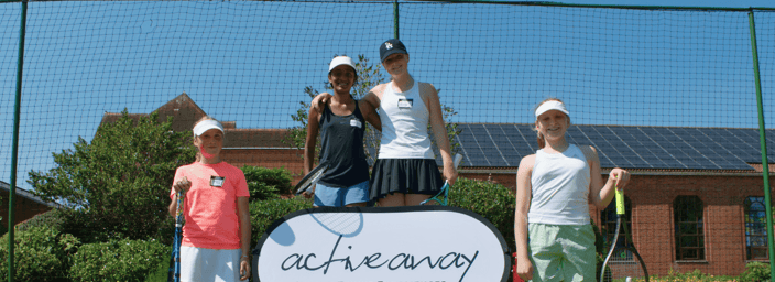 Four junior tennis players smiling on a grass court with rackets and an Active Away sign.