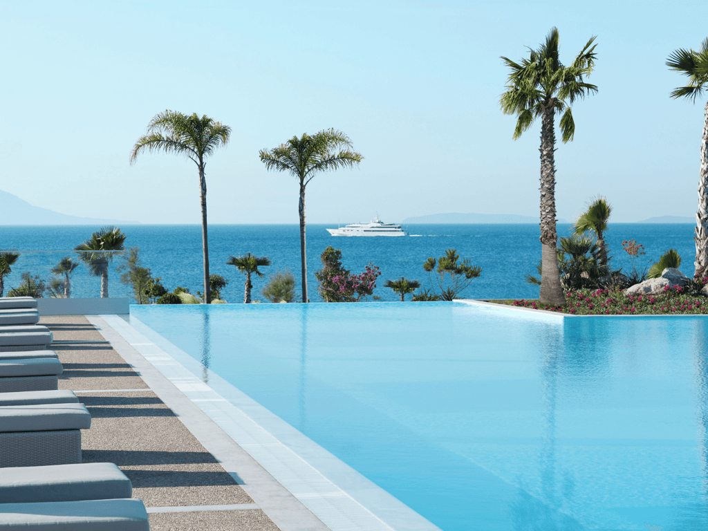Infinity pool with palm trees overlooking the ocean, sun loungers and a yacht in the distance.