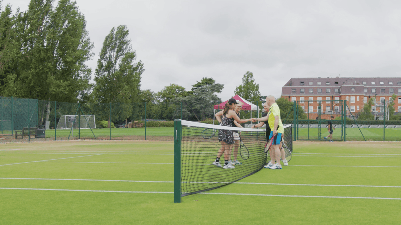 Four tennis players shake hands over a grass-court net with the Lensbury building in the background.