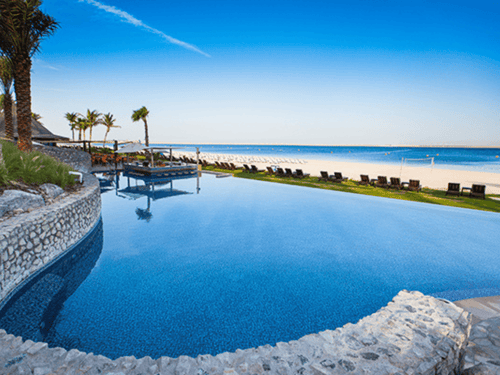 Infinity pool overlooking a sandy beach with palm trees, loungers and calm blue sea at a resort.
