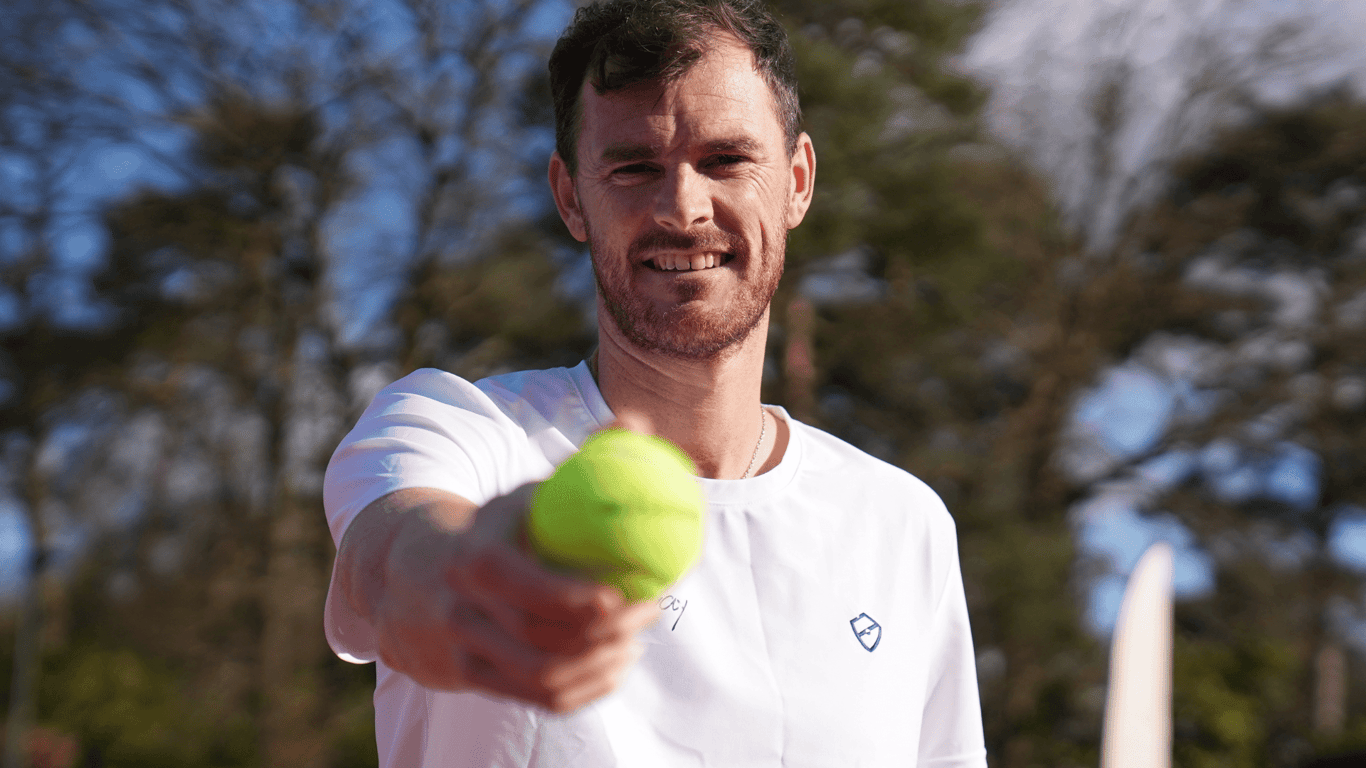 Smiling tennis player holds an Active Away tennis ball toward the camera outdoors.