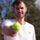 Smiling tennis player holds an Active Away tennis ball toward the camera outdoors.