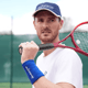 Tennis player in white shirt and cap resting a racquet on his shoulder on an outdoor court.