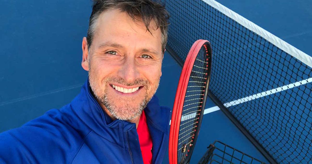 Smiling man with a tennis racket taking a selfie on a blue court beside the net.