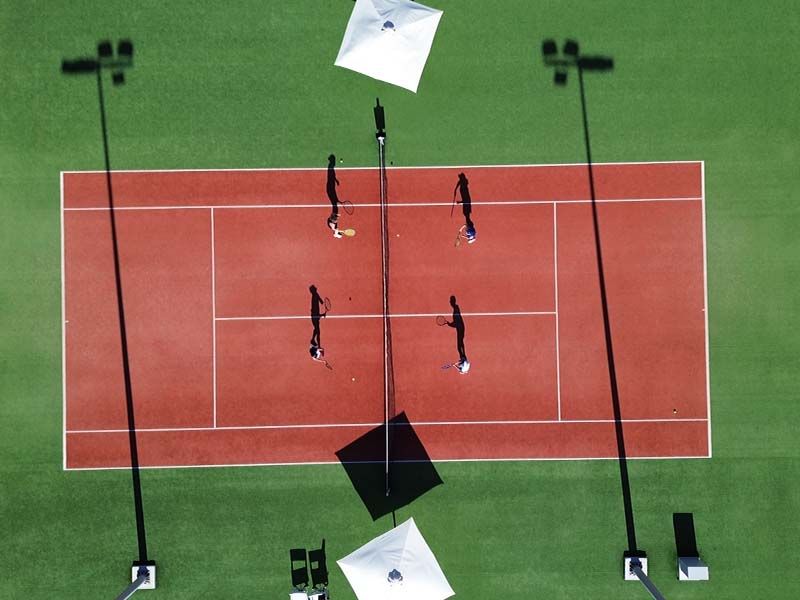 Aerial view of four players playing doubles on a red tennis court with long shadows.