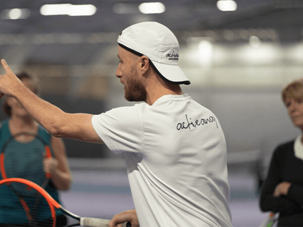 Tennis coach in a white cap gives instructions on an indoor court during an Active Away clinic.