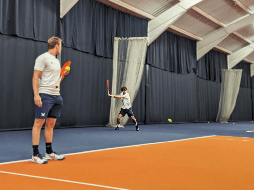Indoor tennis coaching session; coach watches as a player hits a forehand on an orange-and-blue court.