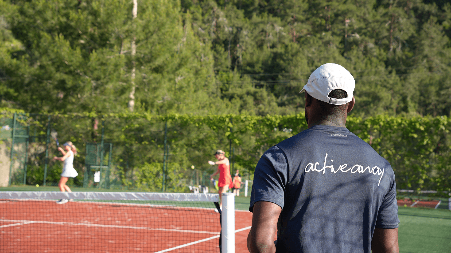 Tennis coach in an Active Away shirt watching players on a sunny outdoor clay court.