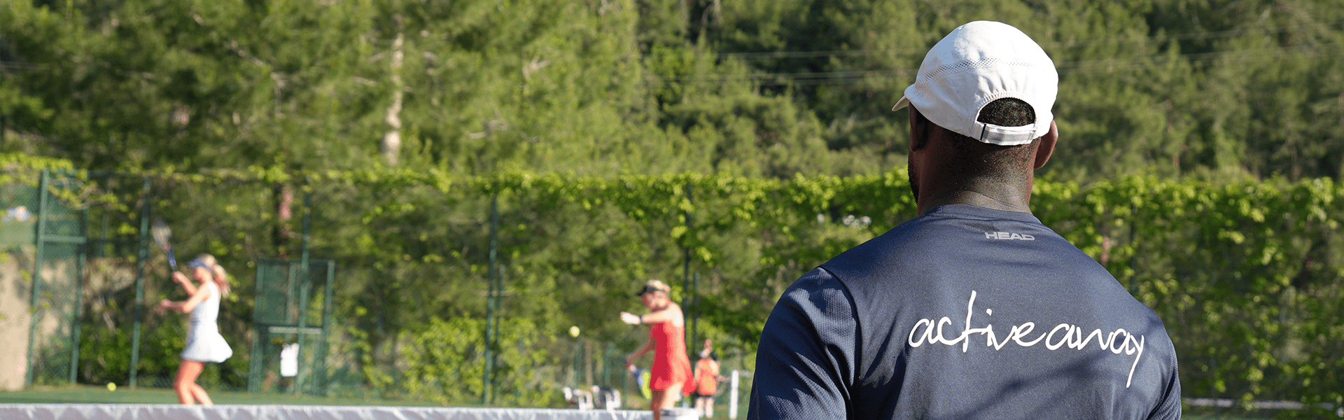 Tennis coach in an Active Away shirt watching players on a sunny outdoor clay court.