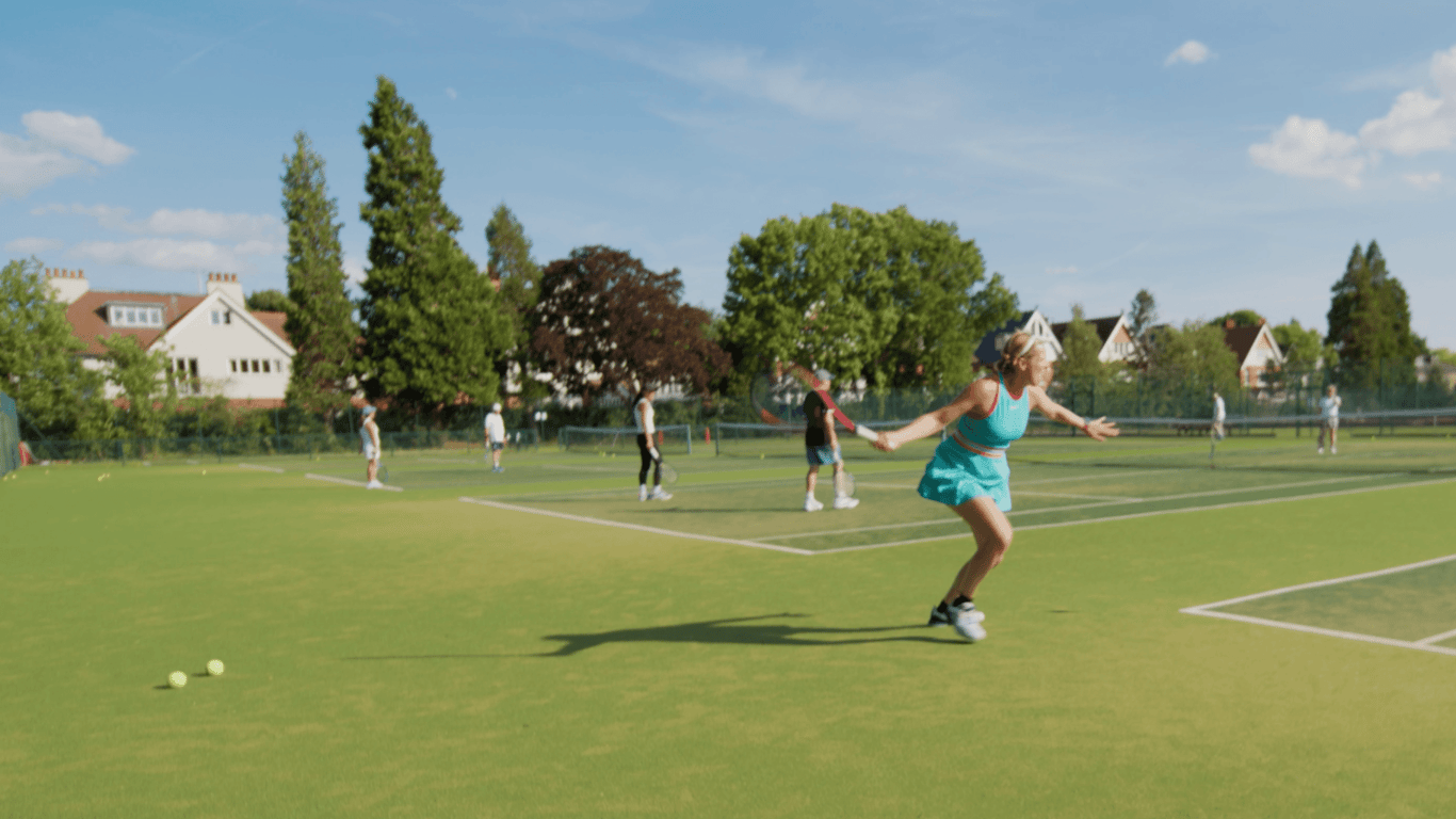 Woman practising tennis footwork on grass courts at Lensbury, with other players training on a sunny day.