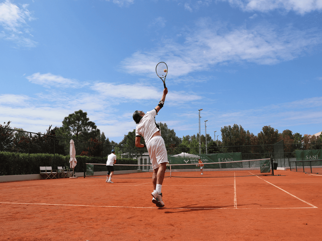 Tennis player serves on a clay court at Sani Beach, with partner ready under a bright blue sky.