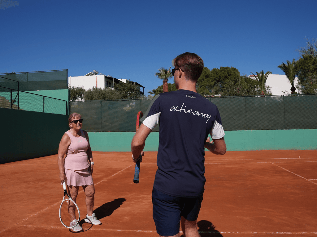 Tennis coach in Active Away shirt instructs a player on a sunny clay court.