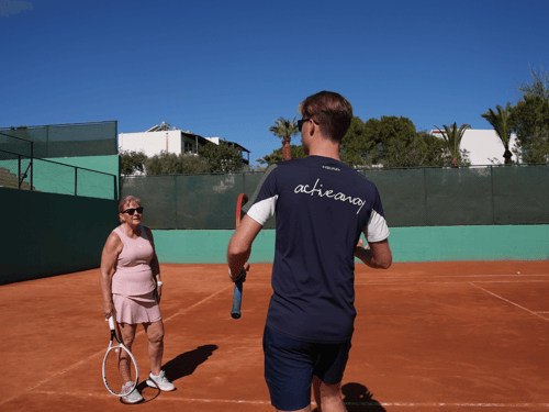 Tennis coach in Active Away shirt instructs a player on a sunny clay court.