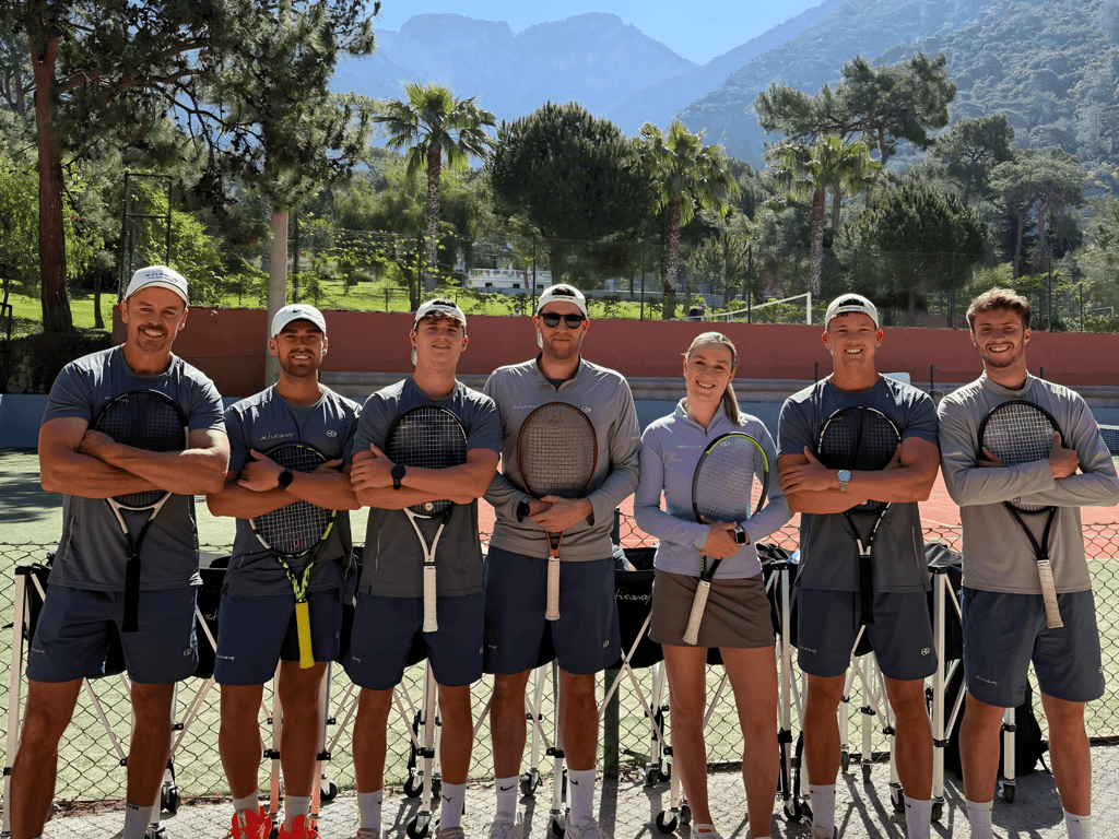 Seven tennis coaches posing with rackets on an outdoor court, mountains and palm trees in the background.