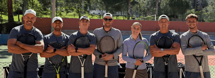 Seven tennis coaches posing with rackets on an outdoor court, mountains and palm trees in the background.