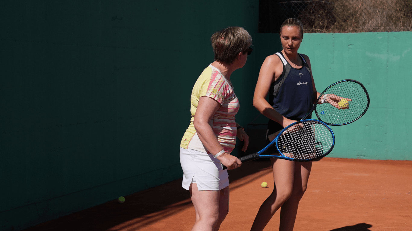 Two women with tennis racquets talk on a clay court during a coaching session.