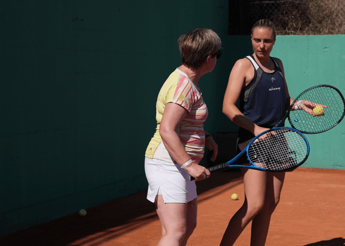 Two women with tennis racquets talk on a clay court during a coaching session.