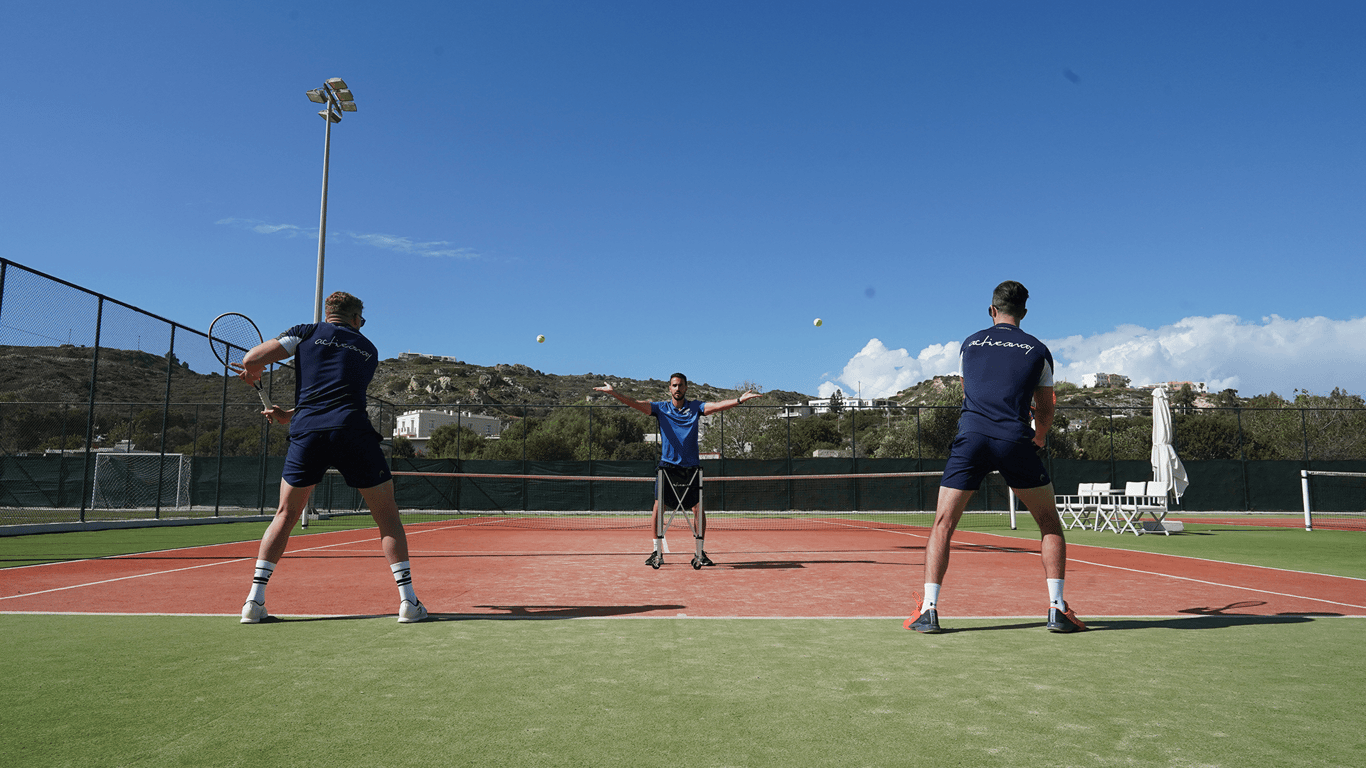 Two tennis players practise as a coach feeds balls on an outdoor court under a clear blue sky.