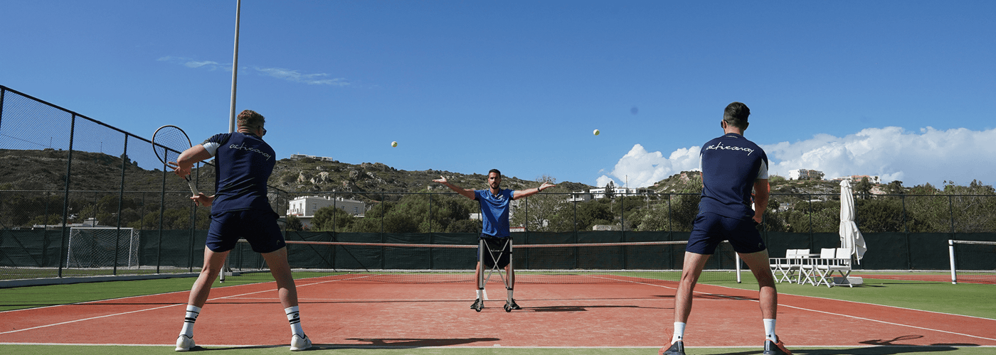 Two tennis players practise as a coach feeds balls on an outdoor court under a clear blue sky.