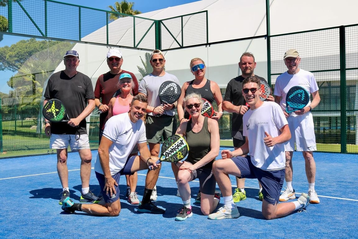 Smiling group holding padel rackets on a blue outdoor court during a sunny coaching holiday.
