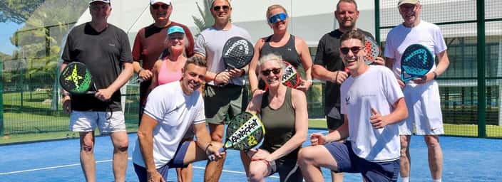 Smiling group holding padel rackets on a blue outdoor court during a sunny coaching holiday.
